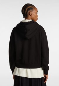 Haut à capuche noir court avec une grande capuche et une coupe ample, présentant des poignets et un ourlet côtelés. Porté par-dessus une chemise blanche à rayures avec des bords effilochés.