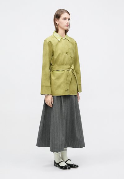 Νεαρή γυναίκα που φοράει ανοιχτό πράσινο μπουφάν με ζώνη, μακριά γκρι φούστα, λευκές κάλτσες και μαύρα πάνινα παπούτσια με ασημένιες τρύπες, στέκεται πλαγίως.