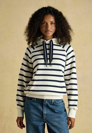 Kvinde med krøllet hår iført marineblå og hvidstribet hættetrøje og blå jeans mod beige baggrund, stående med afslappet kropsholdning.