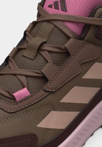 Κοντινό πλάνο καφέ και ροζ sneaker Adidas με αερόβια ύφασμα, ραμμένες συνθετικές επικαλύψεις, σταυρωτά κορδόνια και λογότυπο στη γλώσσα του παπουτσιού.