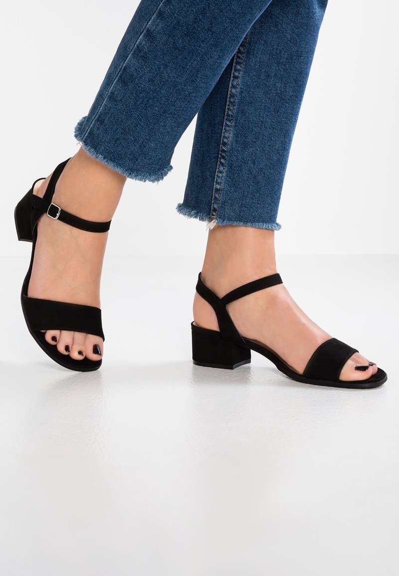 Gros plan sur des pieds portant des sandales noires à talon bloc bas et un jean bleu à ourlet effiloché sur un sol blanc.