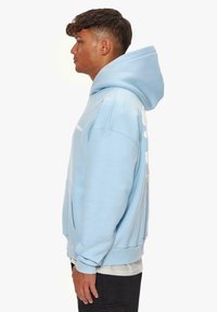 Helleblaue Kapuzenjacke aus softem Stoff, ausgestattet mit einer großen Kapuze, einer vorderen Tasche und einem weißen Logodetail auf der Rückseite. Klassischer Schnitt mit gerippten Bündchen.