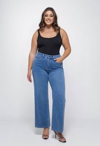 Vrouw met golvend haar, gekleed in een zwarte tanktop, hoge taille brede blauwe jeans en zwarte hoge hakken, staand en lachend.