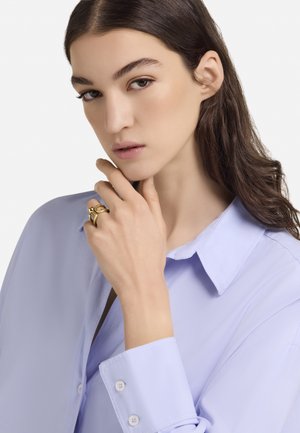 Lyse lilla skjorte med knapper og bred krave. En model bærer en guldring på sin højre hånd med en glat tekstur og abstrakt design.