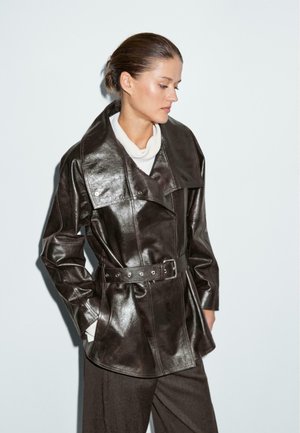 Dark brown leather jacket with a wide collar, belted waist, and button details, featuring a glossy texture. Model wears a white top underneath.
