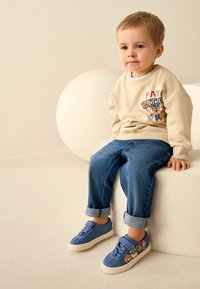 Beżowy sweter z grafiką kreskówkową, niebieskie dżinsy z podwiniętymi nogawkami oraz niebieskie sneakersy z dekoracyjnymi detalami. Siedzący na jasnej powierzchni.
