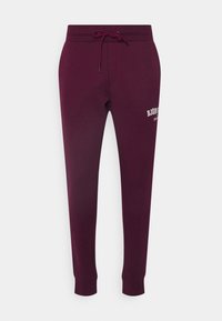 Burgundy svettbyxor tillverkade av en bomullsblandning, med midjeband med snörning, ribbade muddar och tryckt textlogga på vänster lår.