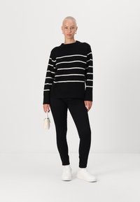 Zwart-wit gestreepte trui met ronde hals, gecombineerd met zwarte leggings en witte sneakers; houdt een kleine witte handtas vast.
