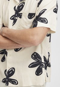 Rövid ujjú, krém színű ing, nagy fekete virágmintákkal, elülső zsebbel és gombokkal, könnyű anyagból készült.
