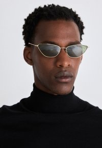 Ochelari de soare stil cat-eye cu ramă aurie și lentile întunecate, având un design elegant. Purtați cu un pulover negru cu guler înalt.