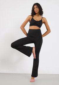 Zwarte cropped top met een twist aan de voorkant, gecombineerd met zwarte flaired broek. Het model draagt roze sokken. Gladde stof, aangesloten ontwerp.