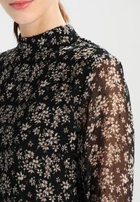 Blouse noire à fleurs avec un col montant, des manches longues transparentes, ornée de petites fleurs blanches et roses, et de délicats accents métalliques.