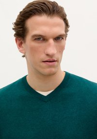 Teal V-hals trui, gebreid materiaal, geribbelde halslijn, gelaagd over een wit shirt. Minimalistisch ontwerp met een klein logo detail op de borst.