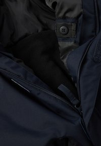 Nahaufnahme einer marineblauen Jacke mit teilweise geöffnetem Reißverschluss, schwarzem Fleece-Futter und schwarzem Knopf an einem Riemen im Kragenbereich.