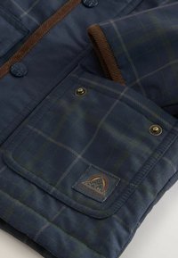 Marineblå quiltet jakke med et grønt ternet mønster, brune corduroy-detaljer og metalliske knapper. Inkluderer et mærket patch på lommen.