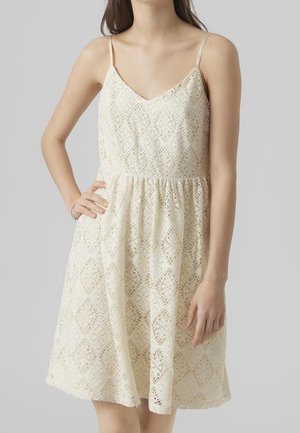 Kvinde iført ærmeløs cremefarvet blondekjole med V-hals og indsnævret talje, stående foran ensfarvet grå baggrund.