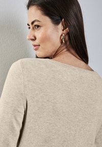 Beige Langarmoberteil mit weitem Ausschnitt, strukturiertem Stoff und gerippten Details. Goldene Creolenakzente ergänzen das Aussehen des Modells.