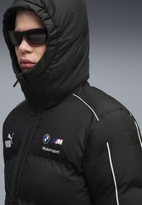 Sort pufferjakke med høj krave og hætte, med hvide accenter, BMW- og Motorsport-logoer, samt let, quiltet tekstur.