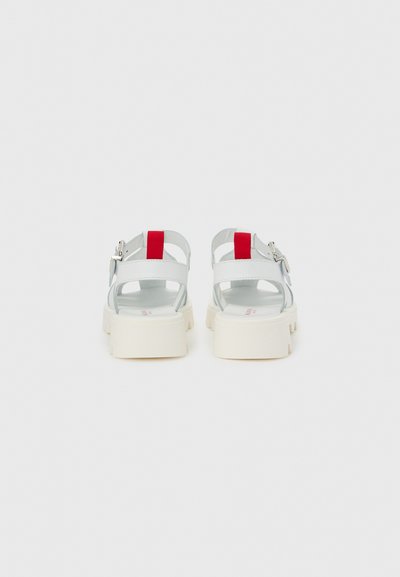 Λευκά πλατφόρμα σανδάλια με κόκκινες λαβές έλξης και ασημένιες λουριά με αγκράφα, φαίνονται από πίσω σε γκρι ανοιχτό φόντο.