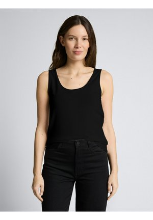 Frau mit langen braunen Haaren, die ein ärmelloses schwarzes Oberteil und schwarze Hosen trägt, steht vor einem schlichten hellgrauen Hintergrund.