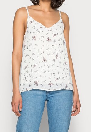 Femme portant un haut blanc sans manches avec des bretelles fines et de petits motifs floraux, associé à un jean bleu clair.