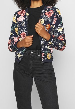 Kvinde iført en marineblå blomstret lynlåstrøje over en sort skjorte og højtaljede sorte jeans, der justerer lynlåsen på jakken med begge hænder.