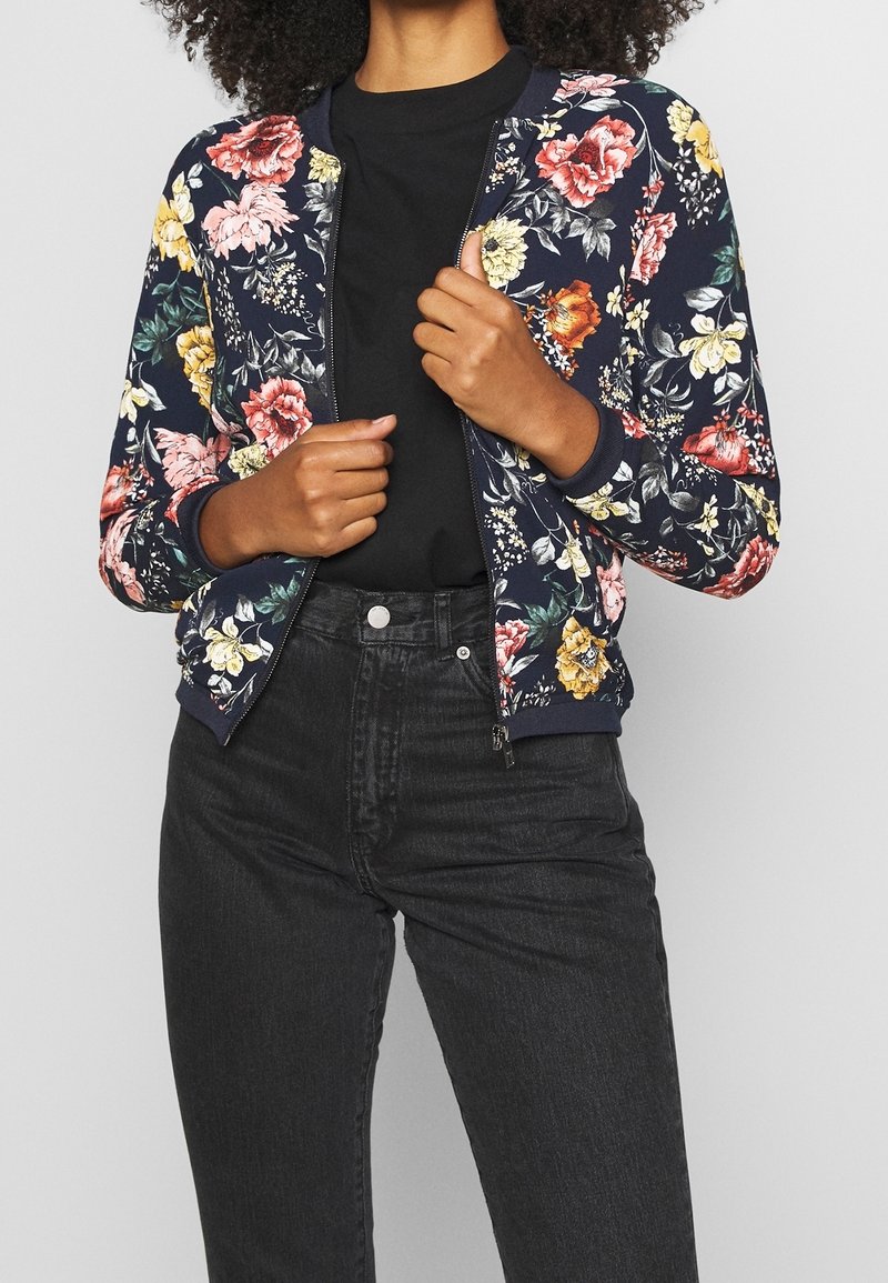 Femme portant une veste zippée bleu marine à motifs floraux sur un t-shirt noir et un jean noir taille haute, ajustant la fermeture éclair de la veste avec ses deux mains.