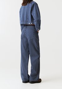 Personne portant un pantalon large à rayures verticales bleu et blanc et une veste courte assortie, debout les mains dans les poches sur un fond blanc.