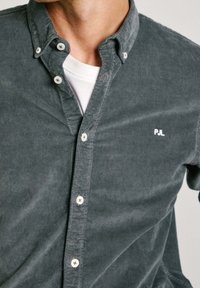 Camicia button-up in corduroy grigio scuro con colletto classico, bottoni bianchi e un piccolo logo bianco sul lato sinistro del petto. Indossata sopra una maglietta bianca.