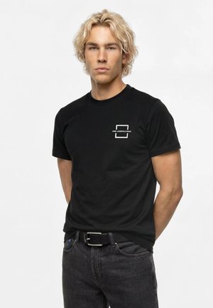 Jeune homme aux cheveux blonds bouclés, portant un t-shirt noir Karl Lagerfeld Jeans et un jean foncé, debout les mains derrière le dos.