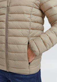 Beige polstret jakke med vandrette quiltede linjer, lynlåslukning foran og sidelommer. Blød, glat tekstur med elastiske manchetter.