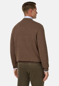 Rudas megztinis su apvalia iškirpte, ilgomis rankovėmis ir laisvu siluetu. Dėvimas ant šviesiai mėlynos marškinių su apykakle ir suderintas su alyvuogių spalvos kordurojo kelnėmis.