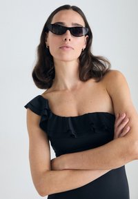 Černé jednodílné plavky na jedno rameno s volánkovým detailem, hladkou texturou a přiléhavým střihem. Tmavé sluneční brýle, vlasy volně upravené.