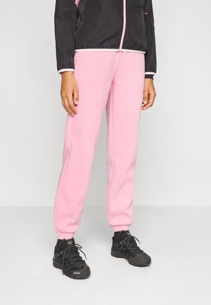 Person trägt hellrosa Sweatpants, schwarze Wanderschuhe und eine schwarze Jacke mit pinkem Reißverschluss und steht auf einem schlichten hellen Boden.