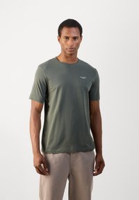 Mörkgrön T-shirt med kort ärm, tillverkad av mjuk bomull. Har en rundad halslinning och liten vit text på vänster bröstområde.