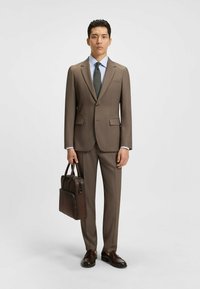 Giacca e pantaloni marroni con una camicia dress azzurra chiara, cravatta a pois e scarpe in pelle marrone. Porta una borsa in pelle marrone.