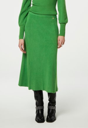 Gebreide groene midi-rok met een aansluitende taille en subtiele ribbelstructuur. Gepaard met zwart kniehoge laarzen met gespikkelde accenten.