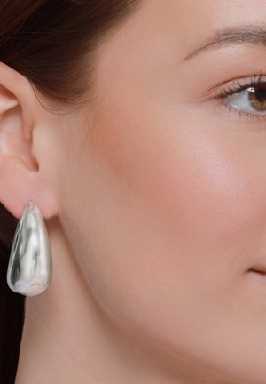 Primo piano del volto di una donna con focus sull’orecchio destro con un grande orecchino argento liscio a forma di goccia.