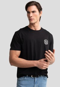 Schwarzes Baumwoll-T-Shirt mit rundem Ausschnitt und kurzen Ärmeln, ausgestattet mit einem weißen runden Logo auf der linken Brustseite und einem lässigen Schnitt.