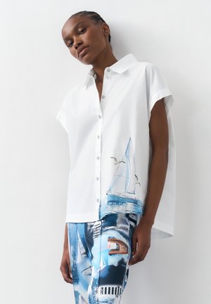 Γυναίκα που φορά λευκό πουκάμισο με κοντά μανίκια και κουμπιά, διακοσμημένο με σχέδια ιστιοφόρου και γλάρου, συνδυασμένο με μπλε παντελόνι με σχέδια ιστιοφόρων και σκαφών.