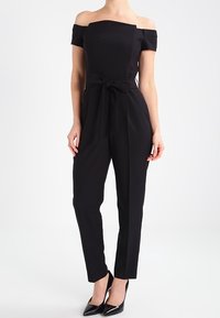 Svart off-shoulder jumpsuit med figurnära bodice, bredt bälte i midjan, smalnade ben och en slät textur. Avslutad med stiliga svarta klackar.
