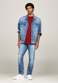 Chaqueta de mezclilla en azul claro con dos bolsillos en el pecho, llevada sobre una camiseta roja. Pantalones vaqueros añiles con detalles desgastados y zapatillas blancas.