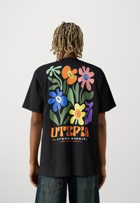 Camiseta de algodón negra con un diseño floral colorido que incluye flores en azul, rosa, naranja y verde. "UTOPIA AVENIR" está impreso en letras en negrita.