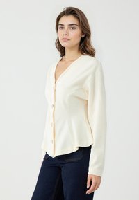 Cremfarbener Cardigan mit V-Ausschnitt, langen Ärmeln und goldenen Knöpfen. Glatte Textur, taillierte Form mit leicht ausgestellt geschnittenem Saum.