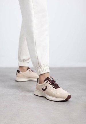 Zapatillas deportivas beige con una textura suave, acentos marrones, cordones en la parte delantera y una suela elevada con un detalle estampado en el lateral.