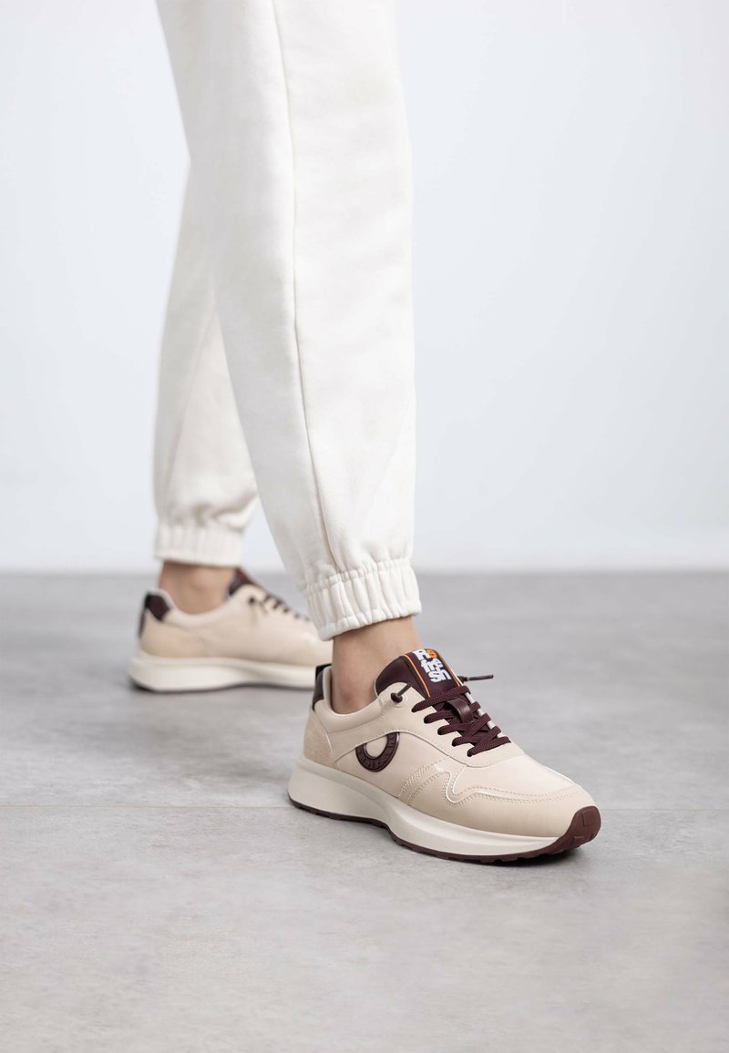 Zapatillas deportivas beige con una textura suave, acentos marrones, cordones en la parte delantera y una suela elevada con un detalle estampado en el lateral.