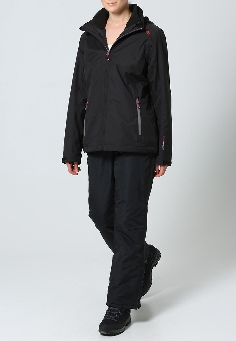 Personne portant une veste imperméable noire avec capuche, un pantalon noir et des chaussures de randonnée noires, debout devant un fond clair uni.