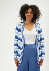 Cardigan a righe blu e bianche con un motivo a zigzag, texture a maglia, apertura frontale e dettagli a coste. Indossato sopra una canotta bianca e pantaloni blu.