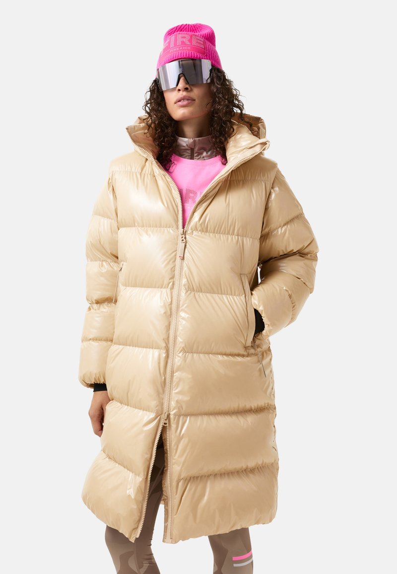 Beige pufferjakke med en blank overflade, der har lynlåslukning, høj krave og oversized lommer. Sammen med en lyserød hue og solbriller.