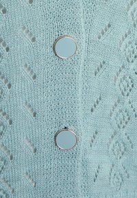 Cárdigan de punto azul claro con un patrón de diamantes, que presenta dos botones metálicos redondos en el lado derecho. Textura suave, grosor medio.